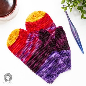 May include: A pair of handmade, knitted mittens in a vibrant gradient of colors. The mittens transition from yellow to red, then to purple and black. The image also shows a cup of coffee, a crochet hook, and a small plant.