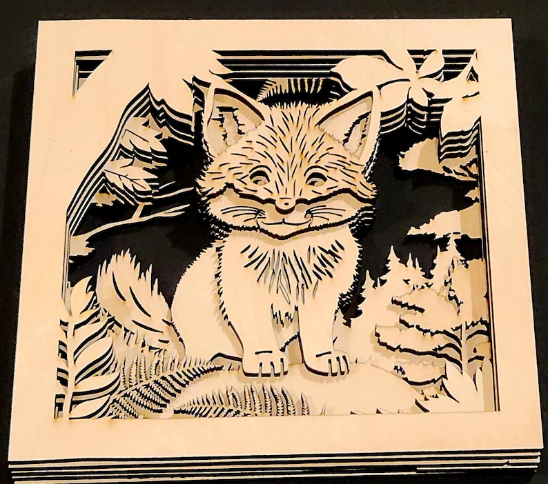 May include: A layered wooden art piece featuring a smiling fox in a forest scene. The fox is light-colored, with detailed fur and facial features. The background includes trees, mountains, and foliage, all cut from wood, creating a 3D effect.