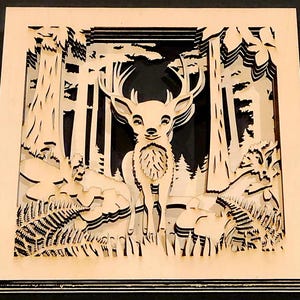May include: A layered wooden art piece depicting a deer in a forest scene. The artwork features a deer with large antlers, surrounded by trees, ferns, and other forest elements. The piece is made of light-colored wood with a dark background.