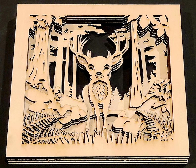 May include: A layered wooden art piece depicting a deer in a forest scene. The artwork features a deer with large antlers, surrounded by trees, ferns, and other forest elements. The piece is made of light-colored wood with a dark background.