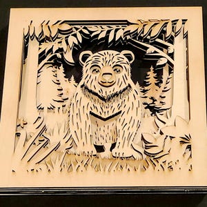 May include: A layered wooden art piece featuring a bear in a forest scene. The bear is the focal point, with detailed fur and a friendly expression. The artwork uses a natural wood color, with a dark background, creating a 3D effect. The piece is square.