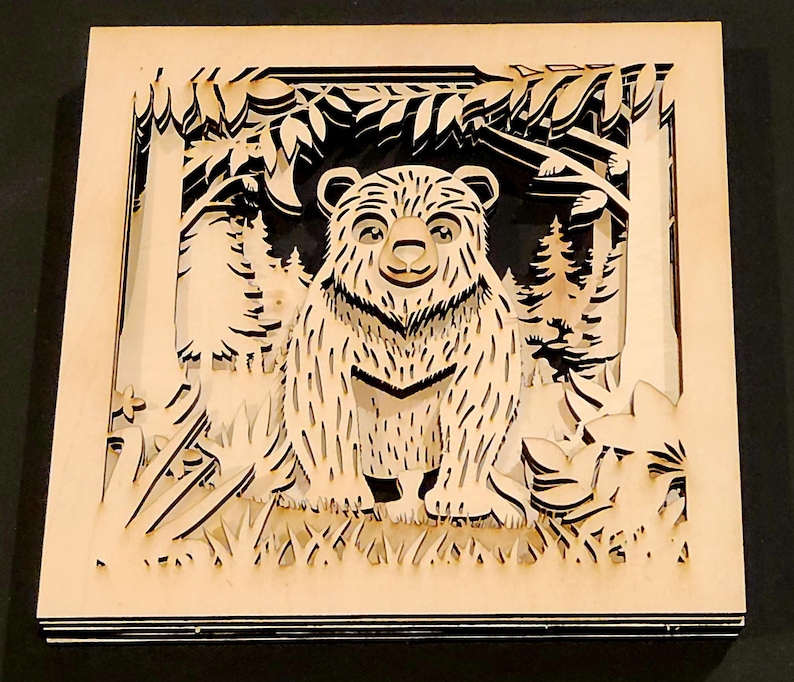 May include: A layered wooden art piece featuring a bear in a forest scene. The bear is the focal point, with detailed fur and a friendly expression. The artwork uses a natural wood color, with a dark background, creating a 3D effect. The piece is square.