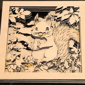 May include: A layered wooden art piece featuring a squirrel holding a nut, surrounded by stylized foliage. The artwork is in a square frame with a light wood finish, and the squirrel and leaves are cut out with black accents.