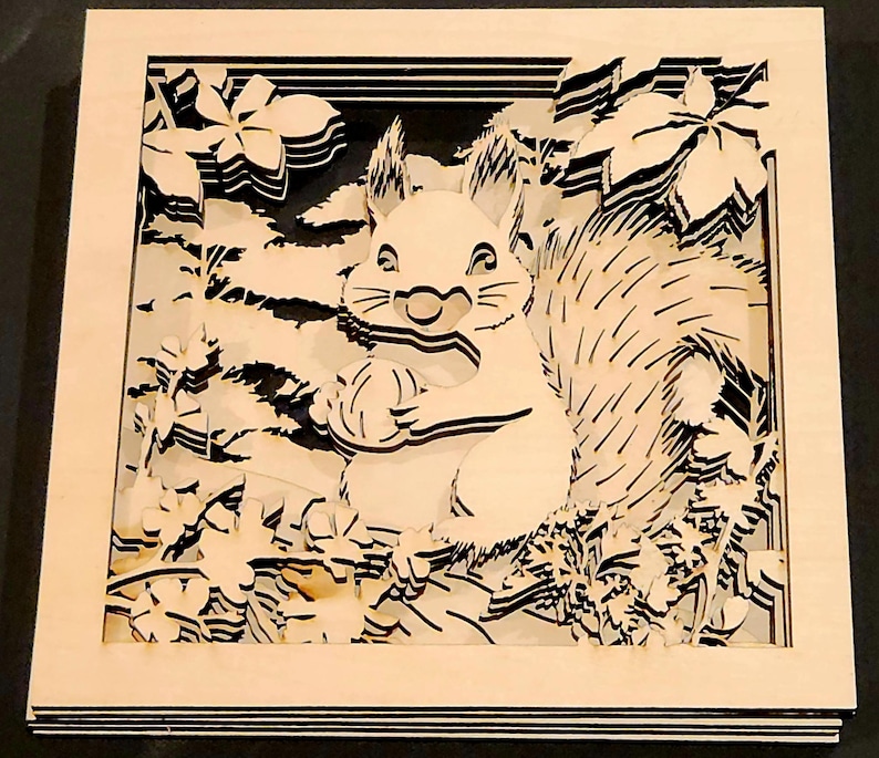 May include: A layered wooden art piece featuring a squirrel holding a nut, surrounded by stylized foliage. The artwork is in a square frame with a light wood finish, and the squirrel and leaves are cut out with black accents.