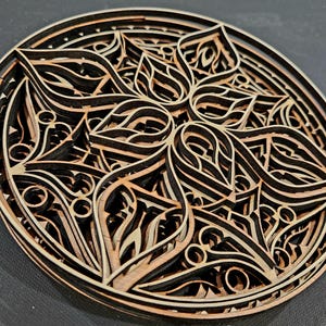 May include: A circular multi-layered wooden wall art piece featuring intricate laser-cut geometric patterns. The design consists of stacked layers of light and dark wood, creating a three-dimensional effect with repeating petal-like shapes.