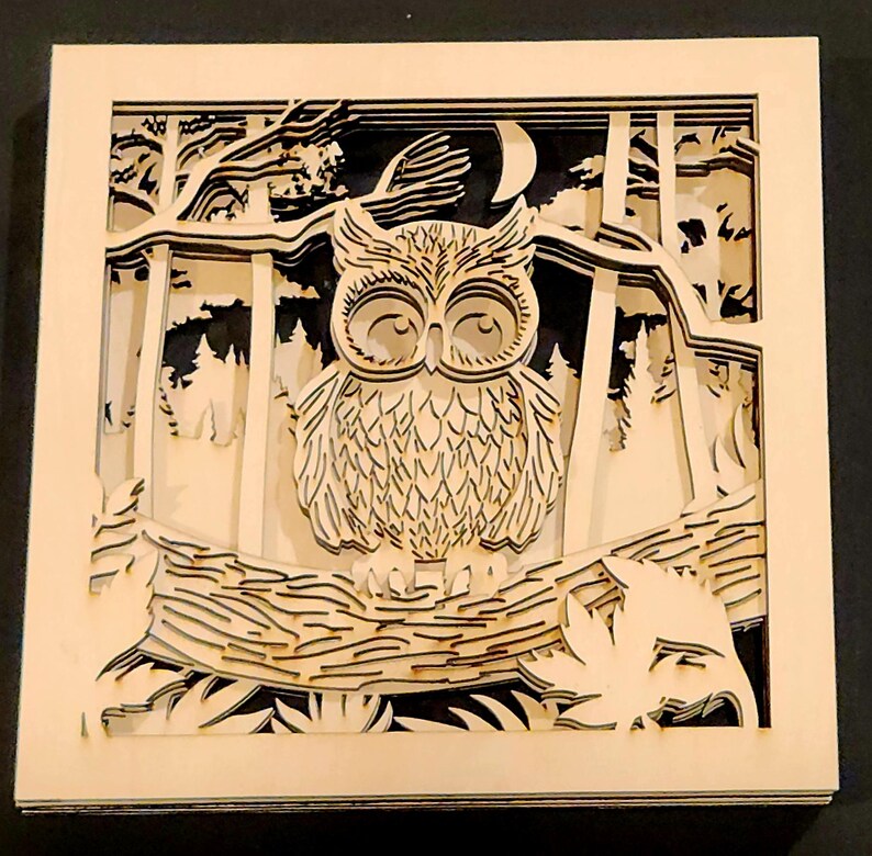 May include: A layered wooden art piece featuring an owl perched on a branch in a forest scene. The artwork is made of light-colored wood, with a crescent moon and trees in the background. The owl has detailed feather patterns.