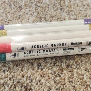 May include: A stack of acrylic markers in a clear plastic wrapper. The markers have white barrels with colored tips in shades of red, green, yellow, and purple. The text "ACRYLIC MARKER" and "Unikniu" are printed on the barrels.