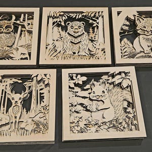 May include: Five square wooden art pieces with layered forest animal designs. Each piece features a different animal: an owl, a bear, a fox, a deer, and a squirrel. The art is in a natural wood colour with black backgrounds.
