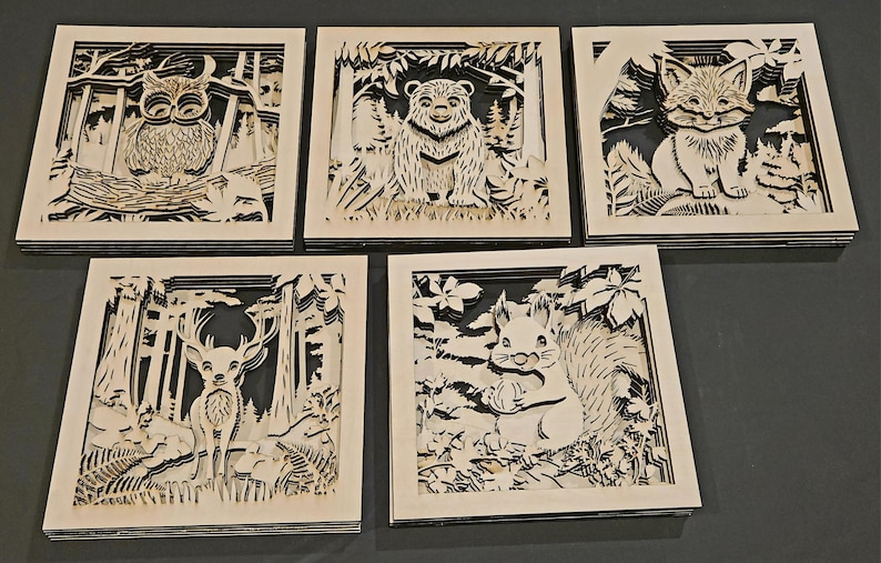 May include: Five square wooden art pieces with layered forest animal designs. Each piece features a different animal: an owl, a bear, a fox, a deer, and a squirrel. The art is in a natural wood color with black backgrounds.