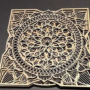 May include: A square, multi-layered wooden mandala with intricate, laser-cut designs. The natural wood tones contrast with the dark spaces created by the cut-out patterns. The design features a central floral motif surrounded by geometric shapes.