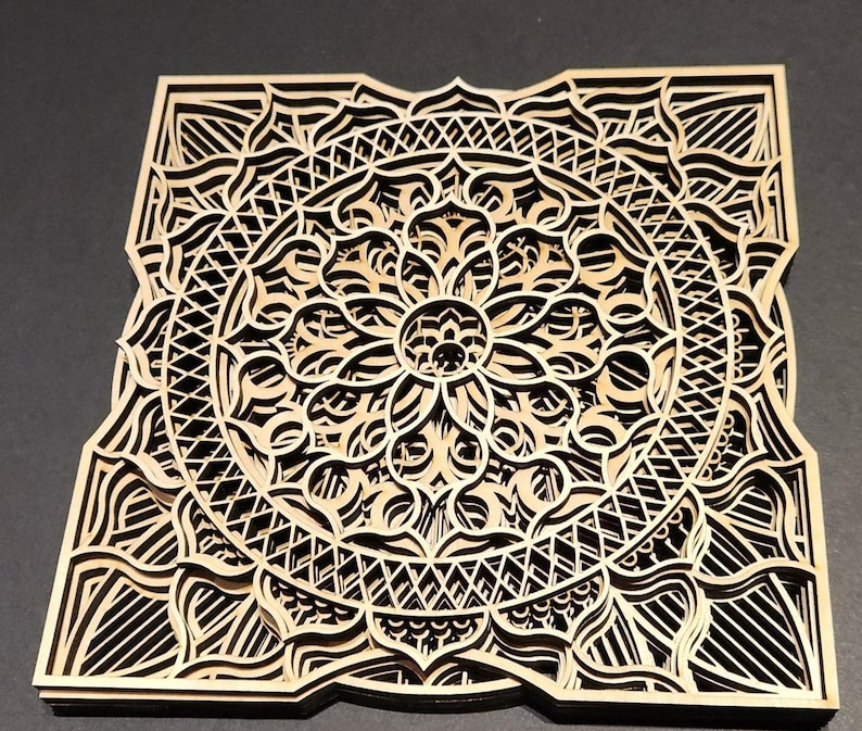 May include: A square, multi-layered wooden mandala with intricate, laser-cut designs. The natural wood tones contrast with the dark spaces created by the cut-out patterns. The design features a central floral motif surrounded by geometric shapes.