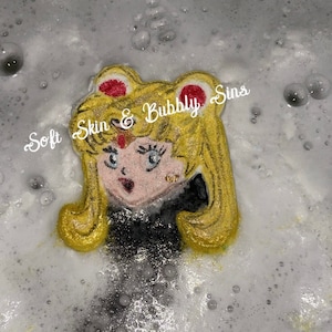 May include: A yellow bath bomb shaped like a cartoon character with a black and white outfit and the text "Soft Skin & Bubbly Sins" on top.