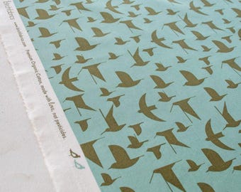 May include: A fabric with a repeating pattern of stylized birds in flight. The birds are a muted gold color against a light blue background. The fabric is organic cotton, made with love, not pesticides.