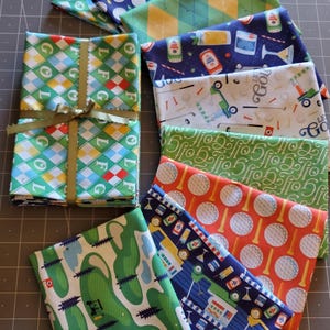 May include: A set of eight fabric squares featuring various golf-themed patterns. The patterns include golf balls, golf carts, golf clubs, and the words "LFG" and "Golf".