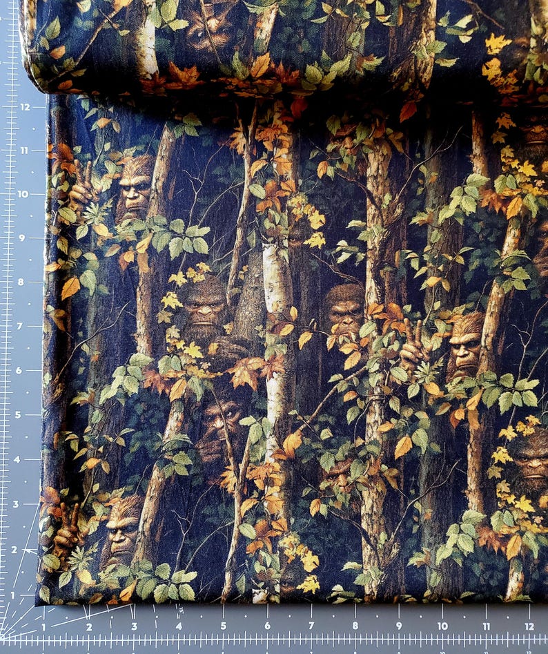 May include: Fabric with a forest scene featuring Bigfoot figures among trees and foliage. The fabric has a dark blue background with brown, green, and yellow leaves. The Bigfoot figures are brown and have a hairy appearance.
