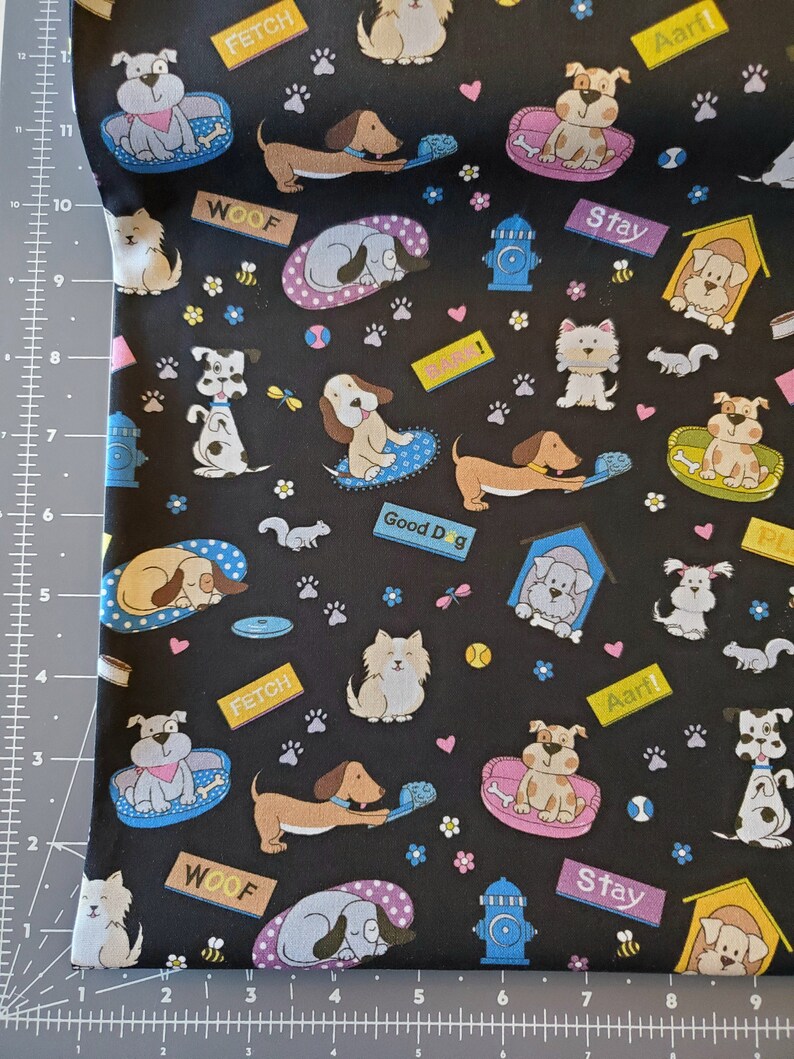 May include: Black fabric with a repeating pattern of dogs in various poses. The dogs are wearing collars and some have bones or toys. The fabric has the words "Woof", "Fetch", "Stay", "Good Dog", and "Aarf!" printed on it.