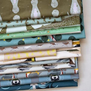 Pode incluir: Uma pilha de vários pedaços de tecido estampado. Os tecidos apresentam desenhos de pássaros, formas abstratas e padrões geométricos em tons de verde, azul, cinza e branco. Os tecidos são dobrados e empilhados, mostrando os diferentes desenhos.
