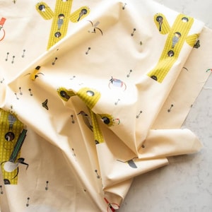 May include: Off-white fabric with a repeating pattern of yellow cacti, birds, and small black arrows. The cacti have black and white striped details. The fabric is folded, showing multiple layers and textures.