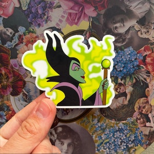 Puede incluir: Una pegatina con Maléfica de La Bella Durmiente de Disney. Se la representa con su piel verde característica, su vestido morado y negro y un bastón verde con un orbe brillante. El fondo es un remolino amarillo y verde.