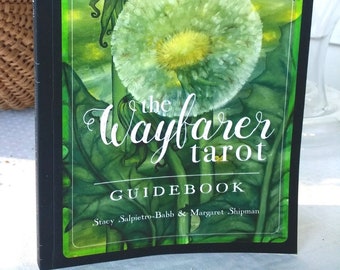 May include: The Wayfarer Tarot Guidebook. The cover shows a dandelion seed head with a blue sky and green field background. The title is in elegant script, with the authors' names below. The book has a black border.