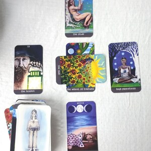 May include: A selection of tarot cards with diverse illustrations. Cards include "The Star," "The Hermit," "The Wheel of Fortune," "High Priestess," "The Moon," and "Queen of Water." The cards are laid out on a white cloth.