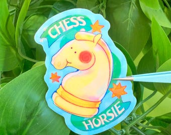 以下が含まれることがあります： 青いバナーに「CHESS HORSIE」の文字が入った、黄色の馬の漫画のチェスの駒のステッカー。 馬は赤い頬をしており、オレンジ色の星に囲まれています。 ステッカーは緑の葉の背景に配置されています。