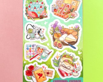 以下が含まれることがあります： 食べ物やオブジェクトのイラストが描かれたステッカーシート。ケーキ、サンドイッチ、カメラ、パレット、本が含まれています。シートは緑色の背景に草のパターンがあり、「Cardinal Cusack @cardinalcrowns」のテキストがあります。