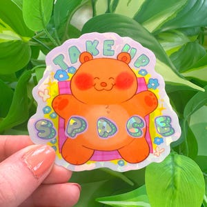 以下が含まれることがあります： ピンク色の頬をしたオレンジ色のクマと、遊び心のあるフォントで「TAKE UP SPACE」という文字が特徴のホログラフィックステッカー。クマは小さな青い花と黄色のアクセントに囲まれています。