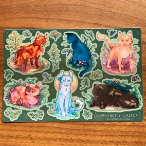 May include: A sticker sheet featuring illustrations of cats in various colors and poses, surrounded by floral and natural elements. The cats include orange, blue, and brown, set against a green background. The artist's name and social media handle are printed at the bottom.