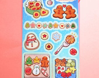 以下が含まれることがあります： 様々なクリスマス用クッキーのステッカーシート。ジンジャーブレッドマン、星、雪だるまなど。クッキーは様々な形と色で、背景は水色で「CARDINAL CUSACK @cardinalcrowns」の文字が入っています。