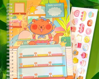 以下が含まれることがあります： カフェをテーマにしたクマのイラストが描かれた、カラフルなスパイラルノート。テキストは「welcome to the STICKER CAFE!」です。様々なペストリーやスイーツのステッカーシートが隣にあります。