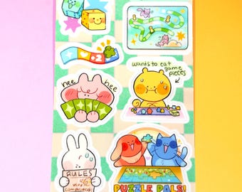 以下が含まれることがあります： ゲームをテーマにした漫画のイラストが描かれた、カラフルなステッカーシート。サイコロ、猫のいるゲーム盤、ゲームの駒を食べたいクマ、そして「RULES very complicated!」と書かれた看板を持ったウサギなどのデザインが含まれています。「PUZZLE PALS!」という文字も見えます。