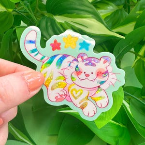 May include: A colorful sticker featuring a cartoon tiger with rainbow stripes and glitter accents. The tiger has a white body, pink cheeks, and a heart-shaped design on its leg. The sticker is surrounded by a light green border and star shapes.