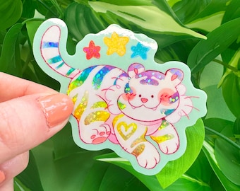 以下が含まれることがあります： 虹色のストライプとキラキラのアクセントが付いた、漫画の虎のカラフルなステッカー。 虎は白い体、ピンクの頬、足にハート型のデザインをしています。 ステッカーは、薄い緑色のボーダーと星の形で囲まれています。