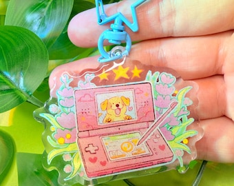 May include: A clear acrylic keychain featuring a pink handheld game console design with a cartoon dog illustration. The keychain has a light blue star-shaped clasp and is decorated with pink flowers and stars.