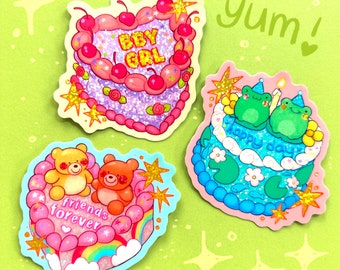 以下が含まれることがあります： ケーキのデザインが施された3つのカラフルなステッカー。1つは「BBY GRL」の文字が入ったハート型、もう1つは2匹のカエルと「happy day」の文字、最後は2匹のテディベアと「friends forever」の文字が入っています。すべてにキラキラとしたアクセントが付いています。