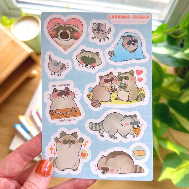 May include: A sheet of raccoon-themed stickers with various illustrations. The stickers feature raccoons in different poses, some with text like "NO THOUGHTS HEAD EMPTY". The background is light blue with a grid pattern, and the text "CARDINAL CUSACK @CARDINALCROWNS" is at the top.