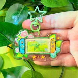 May include: A clear acrylic keychain featuring a video game console design. The keychain has a green star-shaped clasp and a floral border with pink flowers. The game screen shows a character on a horse. The console has blue and orange controllers.