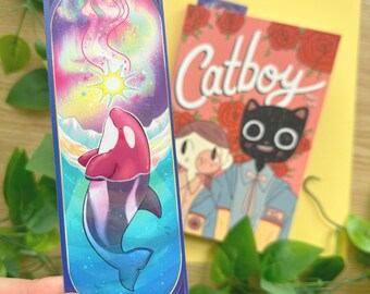 以下が含まれることがあります： ピンクと紫色のシャチのイラストが描かれたブックマーク。カラフルなオーロラを背景にしています。「Catboy」の文字がバラのイラストが入ったピンク色の背景にあります。