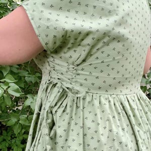 Op de afbeelding: Lichtgroene jurk met een bloemenpatroon en een rug in korsetstijl. De jurk heeft korte mouwen en een volle rok. De stof is een zacht, lichtgewicht materiaal, ideaal voor warm weer. De jurk is in vintage- of cottagecore-stijl.