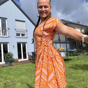 Op de afbeelding: Een oranje en roze patroonjurk met korte mouwen en een getailleerde taille. De jurk heeft een wikkelrok en een veterdetail op het lijfje. De vrouw lacht en draagt oorringen.