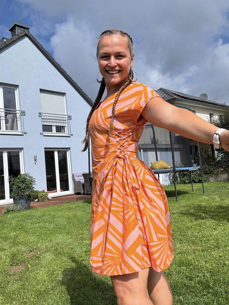 Op de afbeelding: Een oranje en roze patroonjurk met korte mouwen en een getailleerde taille. De jurk heeft een wikkelrok en een veterdetail op het lijfje. De vrouw lacht en draagt oorringen.