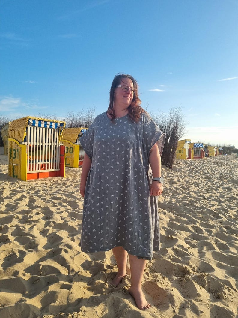 Op de afbeelding: Een grijze, knielange jurk met een wit maansikkepatroon. De jurk heeft korte mouwen en wordt gedragen op een zandstrand. Strandstoelen zijn zichtbaar op de achtergrond onder een heldere blauwe lucht.