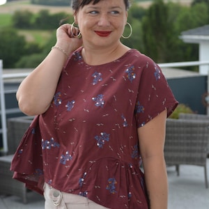 Op de afbeelding: Bordeauxrode top met korte mouwen en een bloemenpatroon in blauw en wit. De top heeft een peplumzoom en wordt gecombineerd met lichtbeige broek. De persoon draagt grote oorringen en een zilveren armband.