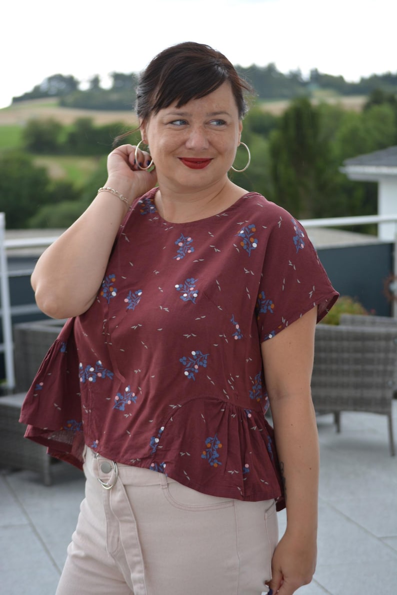 Op de afbeelding: Bordeauxrode top met korte mouwen en een bloemenpatroon in blauw en wit. De top heeft een peplumzoom en wordt gecombineerd met lichtbeige broek. De persoon draagt grote oorringen en een zilveren armband.