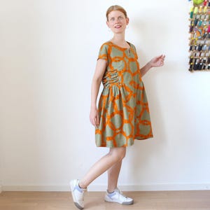 Op de afbeelding: Een olijfgroene jurk met korte mouwen en een herhalend oranje cirkelpatroon. De jurk heeft een geplooid detail aan de zijkant en wordt gedragen met witte sneakers met blauwe accenten.