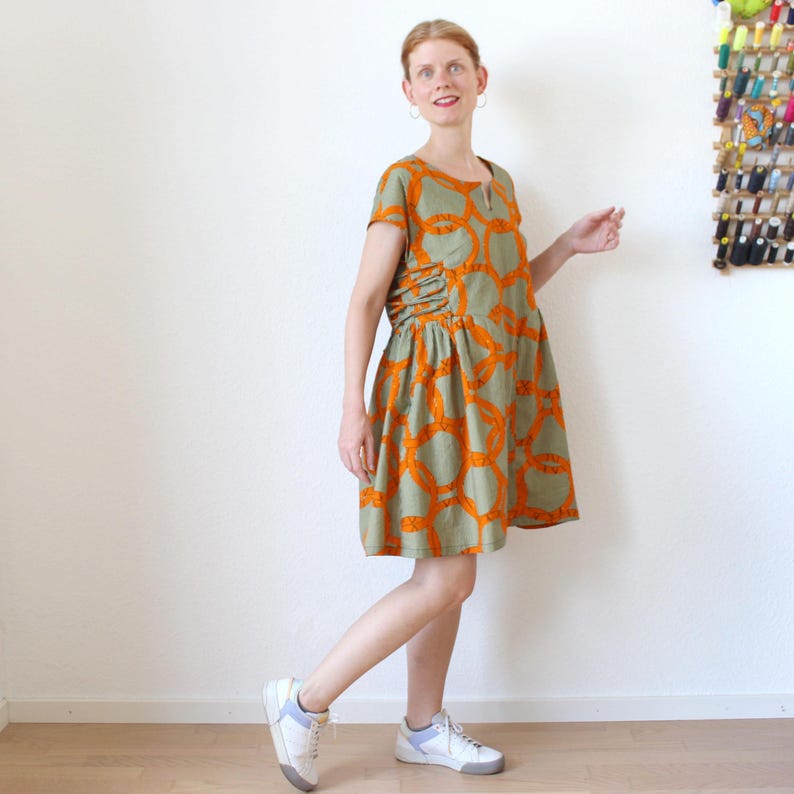 Op de afbeelding: Een olijfgroene jurk met korte mouwen en een herhalend oranje cirkelpatroon. De jurk heeft een geplooid detail aan de zijkant en wordt gedragen met witte sneakers met blauwe accenten.