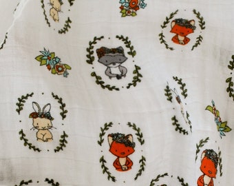 May include: White muslin swaddle blanket with a repeating pattern of foxes, bunnies, and raccoons wearing floral crowns. The animals are surrounded by green wreaths and colorful flowers.