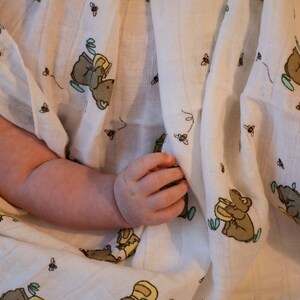 May include: A white muslin swaddle blanket with a repeating pattern of small bees and bears holding honey pots. The blanket is folded and a baby's arm is visible.
