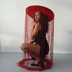 Puede incluir: Un sombrero rojo grande de ala ancha con flecos de cuentas rojas a juego. Los flecos cuelgan, creando un efecto de cortina. El sombrero es el punto focal, con una persona que lleva un vestido negro y tacones negros.